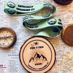 May include: A wooden round plaque with the words "Pen y Fan 886m Officially Smashed It!"  The plaque features a black silhouette of mountains with a dragon at the top. The plaque is sitting on a map of Scotland.