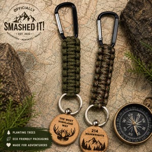 May include: Two keyrings with black carabiner clips and braided cords in olive green and camouflage patterns. Wooden tags feature engraved text and mountain and deer graphics. The text reads "OFFICIALLY SMASHED IT!" and "THE WEST HIGHLAND WAY" and "214 WAINWRIGHTS".