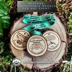 May include: Three circular wooden medals with bark edges, each featuring unique laser-engraved text and graphics. A teal ribbon printed with the text "OFFICIALLY SMASHED IT!" and mountain icons is draped across the medals. The text "BESPOKE, SUSTAINABLE MEDALS FOR ANY EVENT!" and "OFFICIALLYSMASHEDIT.COM" are visible.
