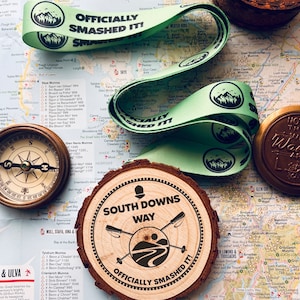 May include: A wooden coaster with the text "SOUTH DOWNS WAY OFFICIALLY SMASHED IT!" sits on a map. Two green ribbons with the text "OFFICIALLY SMASHED IT!" and a compass are also on the map.