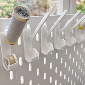 May include: A white pegboard organizer featuring multiple angled hooks and circular cradles designed for holding sewing supplies. The setup includes a spool of grey thread, a small bobbin of beige thread, and a spool of white thread mounted on the hooks.