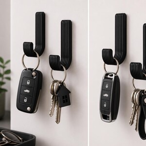 Op de afbeelding: Twee zwarte, J-vormige wandhaken met verticale groeven. Elke haak houdt een set sleutels aan een zilveren sleutelhanger. Eén set bevat een autosleutel, de andere een huissleutel. De haken zijn op een witte muur gemonteerd.
