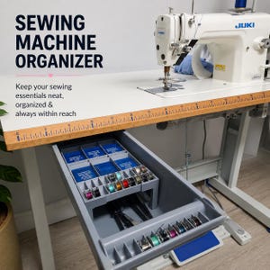 May include: A white Juki sewing machine sits on a table with a built-in wooden ruler measuring up to 115 cm. Below the table, a grey plastic drawer organiser holds blue needle cases, various coloured bobbins, and metal sewing tools.