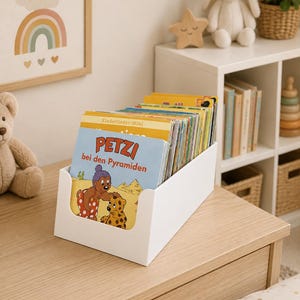 Op de afbeelding: Een witte plastic opbergbak gevuld met een verzameling kinderboeken. Het voorste boek bevat een kleurrijke illustratie van een beer en de tekst "PETZI bei den Pyramiden" met "Kinderlieder-Mini" erboven gedrukt.