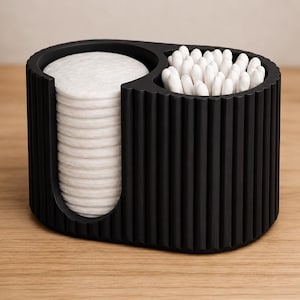 Op de afbeelding: Een zwarte, geribbelde organizer met twee compartimenten. Eén kant bevat een stapel witte wattenschijfjes en de andere kant bevat een verzameling witte wattenstaafjes. De organizer staat op een lichtbruin houten oppervlak.
