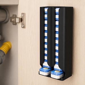 Pode incluir: Um dispensador vertical de parede em preto mate com dois canais paralelos cheios de pastilhas retangulares para máquina de lavar louça empilhadas. Cada pastilha tem uma base branca com uma camada superior azul salpicada, visível pelas ranhuras.