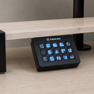 Pode incluir: Um controlador Elgato Stream Deck preto com quinze teclas LCD quadradas com ícones e texto iluminados em azul. O dispositivo está posicionado sobre uma superfície de madeira clara sob um suporte de monitor a condizer.