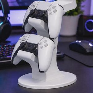 Op de afbeelding: Een witte verticale standaard ontworpen om twee gamecontrollers vast te houden. De standaard heeft een ronde voet en twee trapsgewijze houders, die elk een witte en zwarte draadloze controller ondersteunen met twee analoge sticks en knoppen.