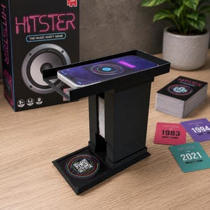 May include: A black Hitster music party game set including a tall card holder stand, a box with a speaker graphic, a deck of cards, and individual game cards labelled 1983, 1994, and 2021. A smartphone rests on the top platform of the stand.