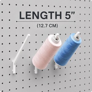 Op de afbeelding: Drie witte, schuine pegboard-haken gemonteerd op een grijs pegboard. Twee haken houden klossen garen vast, één in lichtroze en één in blauw. De tekst LENGTH 5" (12,7 CM) staat boven de haken en geeft de lengte aan.