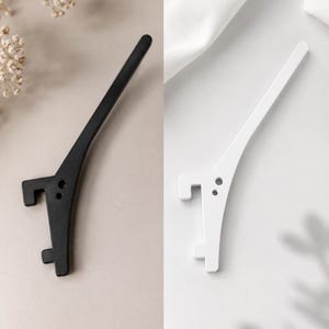 May include: A set of two elongated, curved hooks, one in matte black and one in matte white. Each hook features a long, tapered arm, a flat mounting base with two circular screw holes, and two distinct, squared-off prongs extending outward.