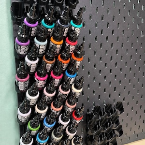 Op de afbeelding: Een zwart geperforeerd wandpaneel met meerdere rijen zwarte plastic flessenhouders. De houders bevatten diverse kleine knijpflesjes Pro Acryl-verf in een breed kleurenspectrum, waaronder paars, turquoise, oranje, rood en wit.