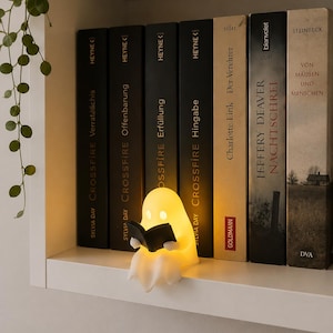 Könnte beinhalten: Eine kleine, leuchtende, weiße Lampe in Geisterform, die auf einem Regal steht. Die Geisterfigur besteht aus einem durchscheinenden Material, strahlt ein warmes gelbes Licht aus und hält ein kleines schwarzes Buch in den Händen vor mehreren Büchern.