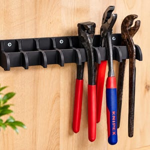 Op de afbeelding: Een zwarte kunststof wandhouder voor gereedschap met meerdere sleuven voor het ophangen van tangen. De houder is bevestigd met schroeven en bevat drie tangen, waaronder één met rood-blauwe handgrepen met het opschrift Knipex en twee roestige metalen tangen.