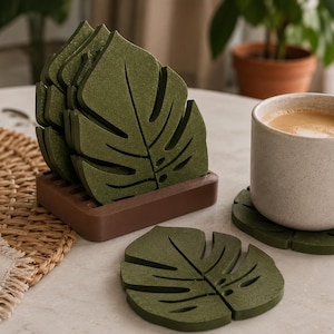 May include: A set of green leaf-shaped coasters made of a textured material. The coasters are in a brown holder, with one coaster next to a mug of coffee. The coasters are designed to resemble a monstera leaf.