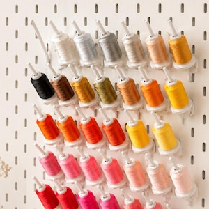 May include: A white pegboard wall organizer holding multiple spools of sewing thread in a gradient of colors including white, gray, brown, yellow, orange, red, and pink, arranged in rows on individual white plastic pegs extending from the board.