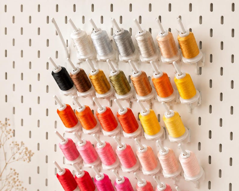 May include: A white pegboard wall organizer holding multiple spools of sewing thread in a gradient of colors including white, gray, brown, yellow, orange, red, and pink, arranged in rows on individual white plastic pegs extending from the board.