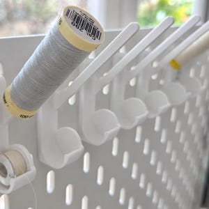 May include: A white pegboard organizer featuring multiple angled hooks designed to hold sewing thread spools. Each hook includes a lower circular cradle for a bobbin. One spool of light gray thread is mounted on the first hook, with a bobbin below.