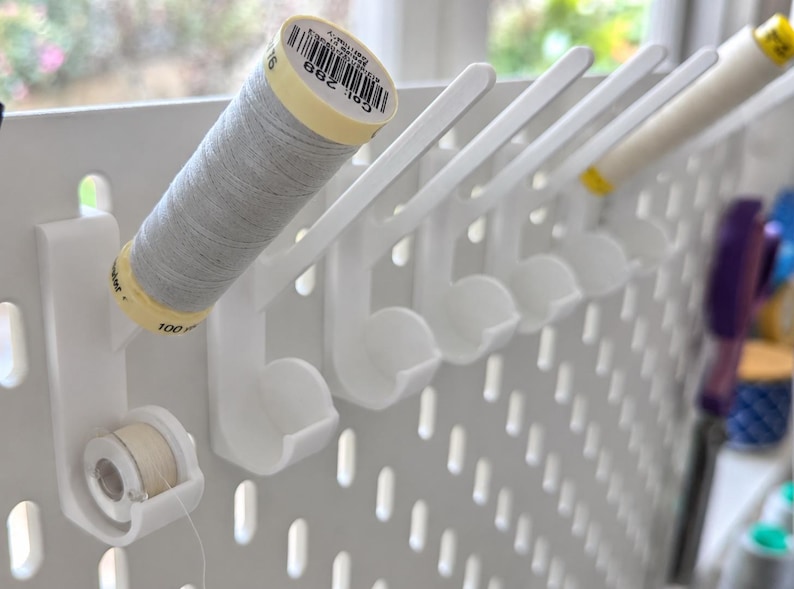 May include: A white pegboard organizer featuring multiple angled hooks designed to hold sewing thread spools. Each hook includes a lower circular cradle for a bobbin. One spool of light gray thread is mounted on the first hook, with a bobbin below.