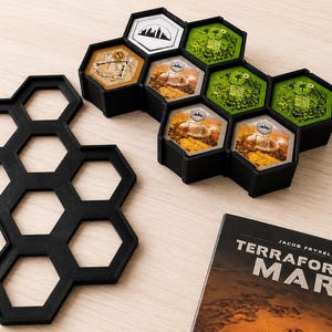 May include: A black plastic hexagonal tile organizer for board games, featuring a grid of seven empty slots. Next to it, a filled organizer holds hexagonal game tiles with green, brown, and white illustrations. A Terraforming Mars game box is visible.