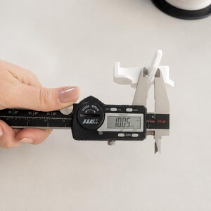 May include: A white plastic component being measured by a black digital caliper. The digital display on the caliper clearly shows a measurement of 10.05 mm. The component features a small, angled structure with a flat base and vertical tabs.