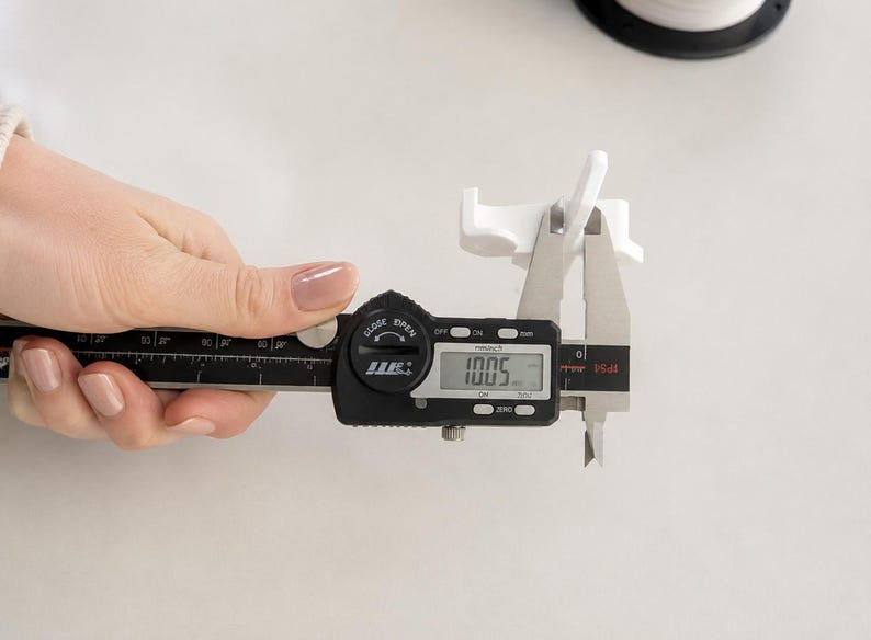May include: A white plastic component being measured by a black digital caliper. The digital display on the caliper clearly shows a measurement of 10.05 mm. The component features a small, angled structure with a flat base and vertical tabs.