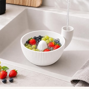 Könnte beinhalten: Eine weiße Obstwaschschüssel aus Kunststoff mit gerippter Außenseite und integriertem seitlichen Ausguss für den Wasserabfluss. Die Schüssel enthält einen zentralen kegelförmigen Einsatz und ist mit frischen roten Erdbeeren, grünen Trauben und Blaubeeren gefüllt.
