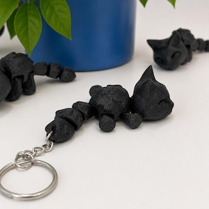 Peut inclure: Porte-clés noir en forme de chat avec un anneau argenté. Le chat est segmenté, permettant le mouvement. Le chat est allongé, les yeux fermés. Un pot bleu avec des feuilles vertes est en arrière-plan.