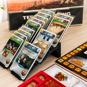 Puede incluir: Un soporte de plástico negro de varios niveles que muestra una serie de cartas de juego de mesa en fundas protectoras. Las cartas presentan diversas ilustraciones, texto y valores numéricos. El soporte está junto a un tablero de juego.