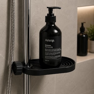 Peut inclure: Une bouteille noire de shampooing Aesop avec un distributeur à pompe est posée sur un porte-savon noir fixé à une barre de douche chromée. La bouteille porte du texte blanc. Le porte-savon est fixé sur un mur carrelé.