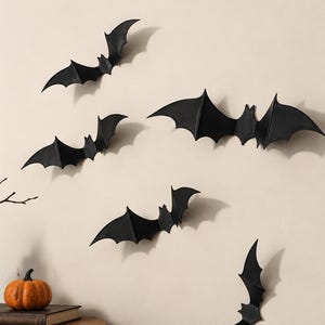 May include: A set of five black bat-shaped wall decorations arranged in a scattered pattern. Each bat features pointed ears, outstretched wings with defined segments, and a central body. The bats are matte black and create shadows on the wall.