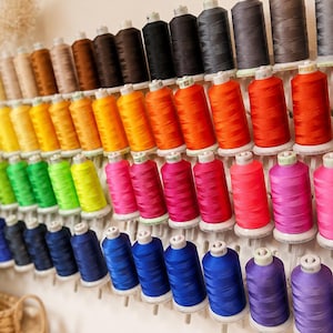 May include: A wall-mounted thread rack holding numerous spools of sewing thread in a wide spectrum of colors, including shades of yellow, orange, red, pink, blue, purple, green, brown, gray, and black, arranged in neat, organized rows.