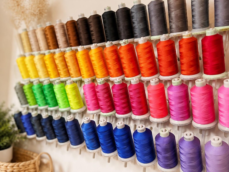 May include: A wall-mounted thread rack holding numerous spools of sewing thread in a wide spectrum of colors, including shades of yellow, orange, red, pink, blue, purple, green, brown, gray, and black, arranged in neat, organized rows.