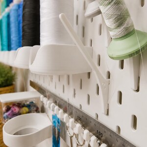 May include: A white pegboard organizer system featuring multiple spool holders, a metal ruler with markings up to 150 cm, and hooks holding metal scissors. The setup includes various colored sewing thread spools arranged in a vertical row.