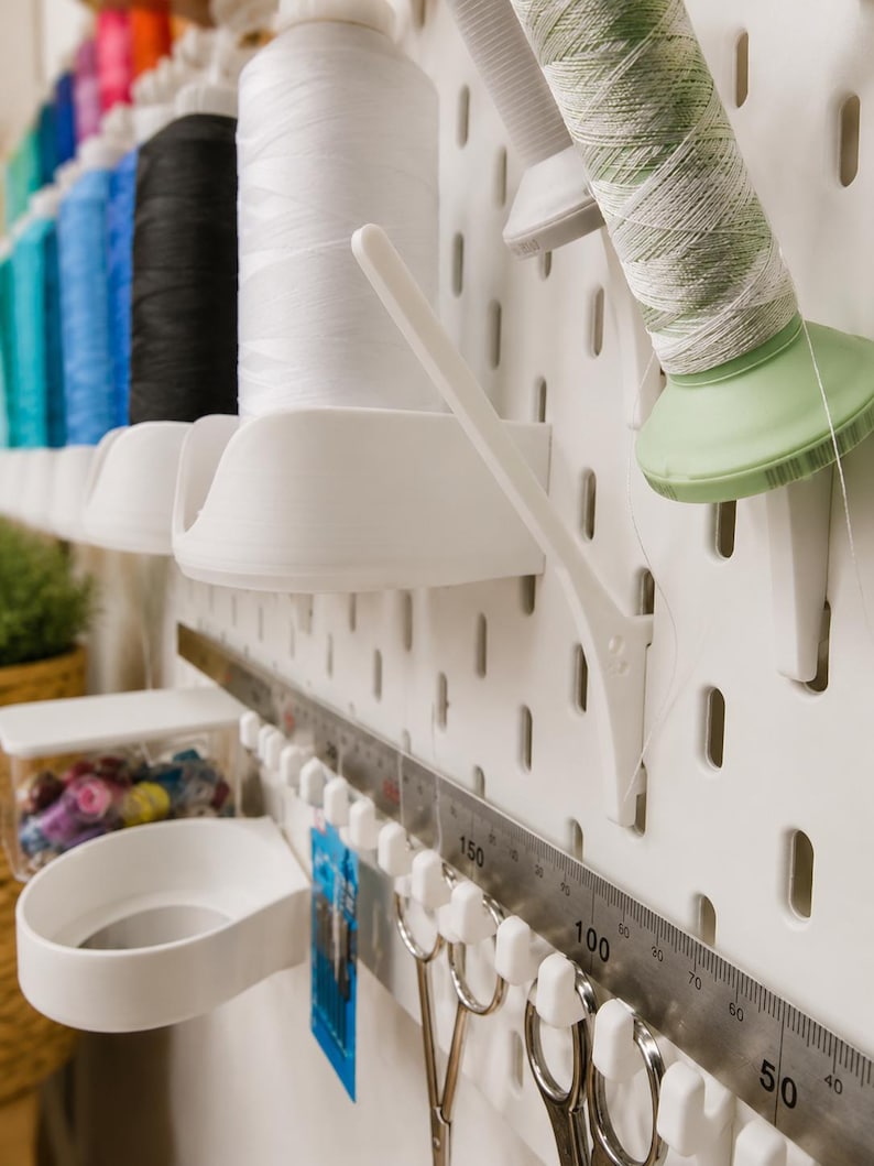 May include: A white pegboard organizer system featuring multiple spool holders, a metal ruler with markings up to 150 cm, and hooks holding metal scissors. The setup includes various colored sewing thread spools arranged in a vertical row.