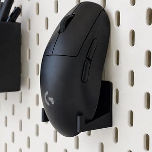 Könnte beinhalten: Eine schwarze kabellose Computermaus mit Scrollrad und Seitentasten, die in einer schwarzen Kunststoffhalterung an einer weißen Lochplatte befestigt ist. Die Maus hat ein graues G-Logo auf der Oberfläche und die Lochplatte zeigt vertikale Schlitze.