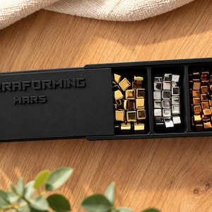 Könnte beinhalten: Eine schwarze, rechteckige Aufbewahrungsbox mit dem eingeprägten Schriftzug TERRAFORMING MARS auf dem Schiebedeckel. Das offene Fach enthält drei Bereiche, die mit kleinen metallischen Würfeln in Gold, Silber und Kupfer gefüllt sind.