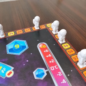 Puede incluir: Un juego de mesa con una superficie de temática espacial oscura y una pista numerada del 43 al 60. Cinco pequeñas figuras de astronautas blancas están colocadas en la pista. Un gráfico de termómetro vertical muestra valores de -4 a +8 y un icono de gota de agua azul muestra el número 9.
