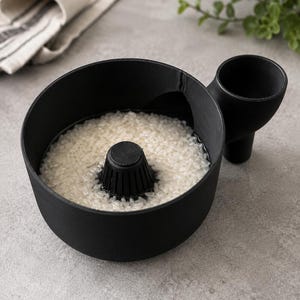 Peut inclure: Un bol à laver le riz circulaire noir mat doté d'un cône de drainage central perforé et d'une poignée latérale avec une ouverture en forme d'entonnoir intégrée. Le bol est rempli de grains de riz blanc crus immergés dans de l'eau.
