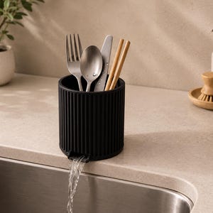 May include: A black cylindrical utensil holder with vertical grooves, holding a fork, spoon, knife, and chopsticks. Water flows from a spout at the base. The holder sits on a countertop next to a sink.