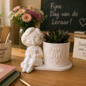 May include: A white ceramic desk organiser featuring a sculpted figure of a person with glasses holding a book next to a cylindrical planter containing a succulent. The planter has the embossed text Beste Juf and a heart symbol on the front.