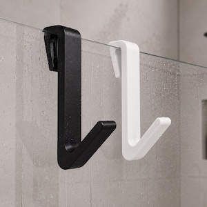 May include: Two shower door hooks, one black and one white, are displayed. The hooks are made of a solid material and are designed to hang over a glass shower door. The hooks have a simple, modern design.