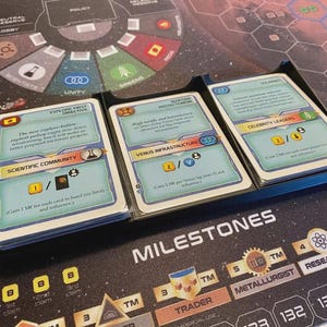 Könnte beinhalten: Drei Stapel rechteckiger Brettspielkarten in einem schwarzen Kunststoff-Organizer. Die Karten haben hellblaue Hintergründe, bunte Symbole und Text, darunter Explore First Directive, Martian Protectionism und Celebrity Leaders.