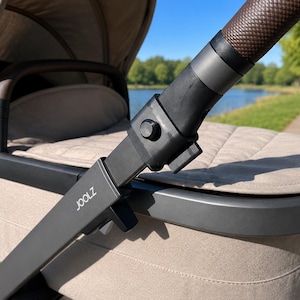 May include: A beige Joolz stroller featuring a quilted fabric bassinet, a black frame, and a dark brown textured leatherette handlebar. The stroller includes a matching beige canopy and a black bumper bar with brown leatherette accents.