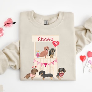 Puede incluir: Una sudadera beige con un gr&aacute;fico de cuatro perros salchicha en un puesto de besos. El puesto est&aacute; decorado con banderines rosas y blancos y el letrero dice "Kisses 5&cent;".