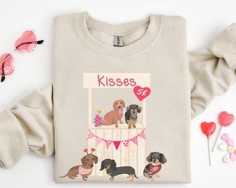 May include: A beige sweatshirt with a graphic of four dachshund dogs at a kissing booth. The booth is decorated with pink and white bunting and the sign says "Kisses 5¢".