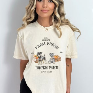 May include: A cream colored t-shirt with a graphic of two miniature schnauzers in front of a pumpkin patch. The text on the shirt reads "The Original Farm Fresh Est'd 1990 Pumpkin Patch Open Daily".