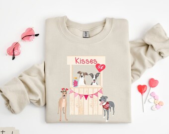 May include: A natural-colored sweatshirt featuring a Valentine's Day themed graphic. The graphic depicts a dog kissing booth with the word "Kisses" above it, and several dogs. The sweatshirt also has the word "natural" printed on it.