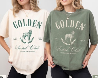Może przedstawiać: Dwie kobiety w dużych koszulkach z grafiką psa i tekstem "GOLDEN Social Club THE HAMPTONS, NEW YORK EST 1983". Koszulka po lewej stronie jest kremowa, a koszulka po prawej stronie jest mchu.