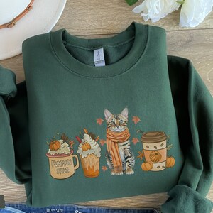 May include: Forest green sweatshirt with a cute cat wearing an orange scarf, surrounded by fall-themed coffee cups with pumpkin spice, whipped cream, and fall leaves. The text "Pumpkin Spice" is on one of the cups.